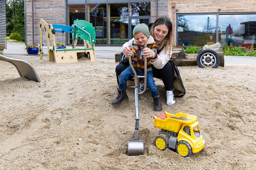 Frau hilft Kind beim Spielen mit Bagger im Sandkasten auf einem Spielplatz