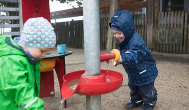 Zwei Kinder spielen draußen mit Wasser an einer roten Wasserspielanlage im Sand