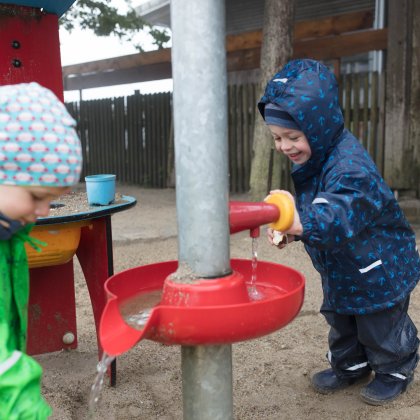 Zwei Kinder spielen draußen mit Wasser an einer roten Wasserspielanlage im Sand