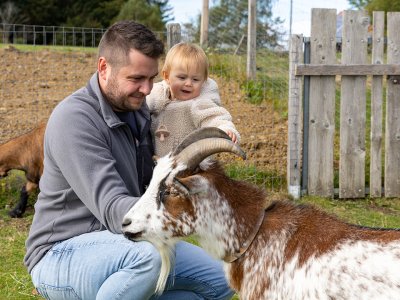 Mann kniet neben Kind, beide streicheln eine Ziege vor einem Holzzaun und Drahtgitter