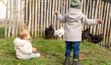 Zwei Kinder in warmer Kleidung vor einem Holzzaun, eines sitzt auf dem Gras, das andere steht mit ausgebreiteten Armen neben Hühnern