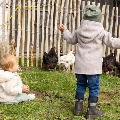 Zwei Kinder in warmer Kleidung vor einem Holzzaun, eines sitzt auf dem Gras, das andere steht mit ausgebreiteten Armen neben Hühnern