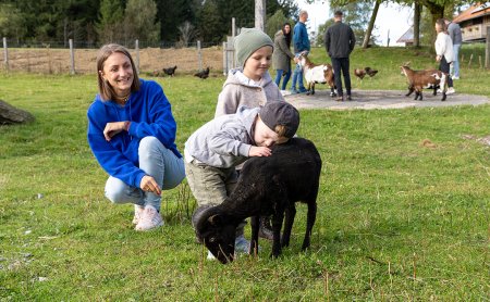 Frau hockt links, zwei Kinder streicheln schwarzes Schaf auf Wiese, im Hintergrund Menschen mit Ziegen