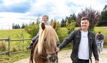 Kind sitzt auf Pferd, Mann führt Pferd, im Hintergrund weitere Personen auf einem Weg im Grünen