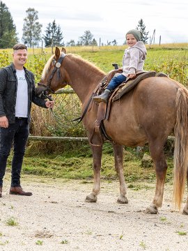 Kind sitzt auf braunem Pferd, Mann hält Zügel, ländliche Umgebung mit Zaun und Bäumen