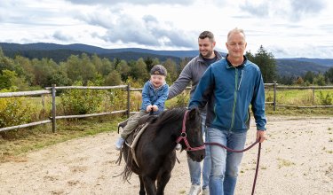 Kind reitet Pony, begleitet von zwei Männern auf einem Weg mit Zaun und Wald im Hintergrund