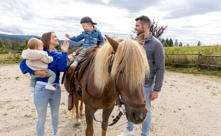 Familie mit zwei Kindern und Pferd auf einem Reitplatz, Kind sitzt auf dem Pferd, Eltern stehen daneben