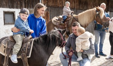 Kinder reiten auf Ponys, begleitet von Erwachsenen vor einem Holzgebäude auf gepflastertem Hof