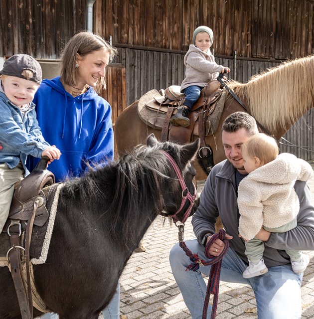 Kinder reiten auf Ponys, begleitet von Erwachsenen vor einem Holzgebäude auf gepflastertem Hof