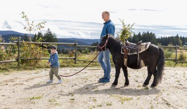Ein Mann hält ein Pony an der Leine, während ein Kind davor läuft auf einem Feldweg mit Zaun und Wald im Hintergrund.