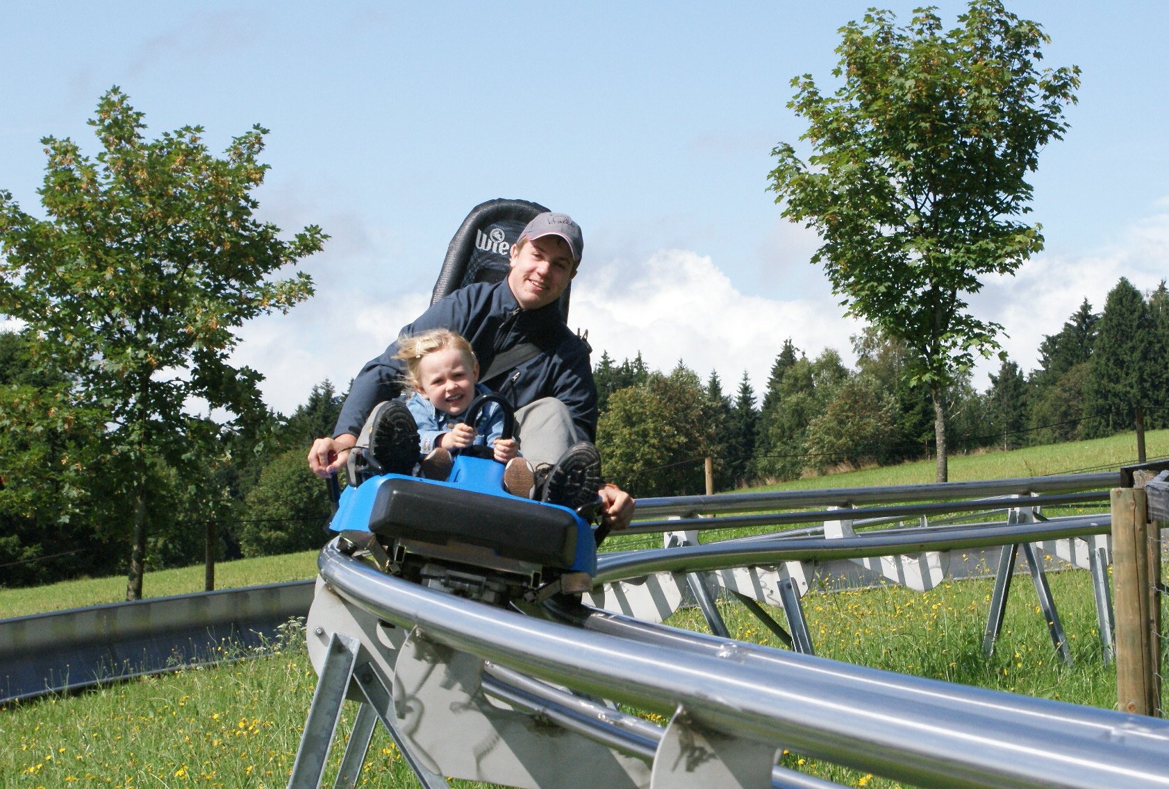 Vater und Kind fahren zusammen eine Sommerrodelbahn auf einer grünen Wiese mit Bäumen