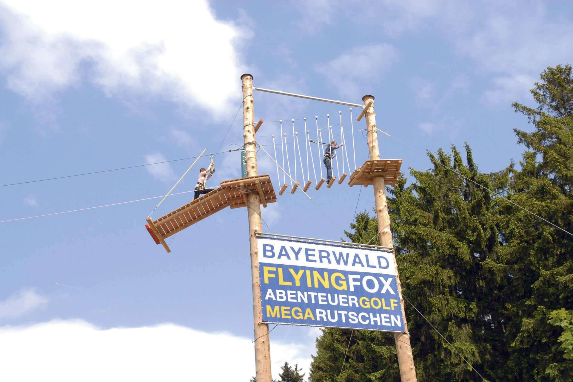 Zwei Personen auf einem Hochseilgarten-Parcours über einem Schild mit der Aufschrift Bayerwald Flying Fox Abenteuer Golf Mega Rutschen