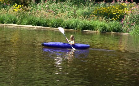 Kind paddelt in blauem Kajak auf ruhigem See vor blühendem Ufer mit grünen Pflanzen