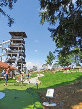 Kinder spielen Minigolf auf grünem Platz neben großem Holzturm mit Rutschen unter blauem Himmel
