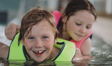Zwei Kinder mit Schwimmwesten liegen im Wasser, eines lächelt in die Kamera