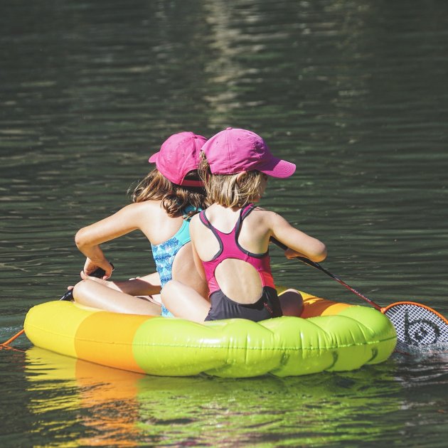 Zwei Kinder mit pinken Mützen sitzen Rücken an Rücken auf einem aufblasbaren Boot im Wasser
