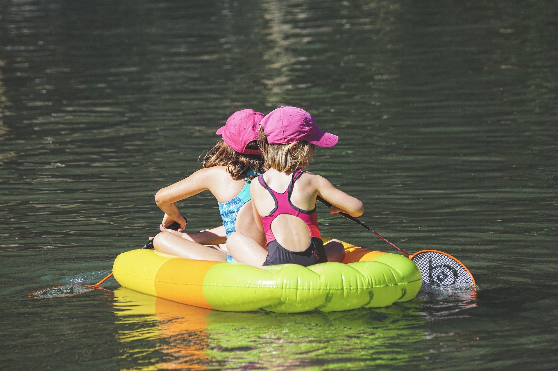 Zwei Kinder mit pinken Mützen sitzen Rücken an Rücken auf einem aufblasbaren Boot im Wasser
