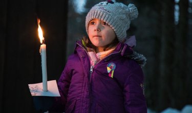 Mädchen in lila Jacke und grauer Mütze hält brennende Kerze im Wald bei Dämmerung