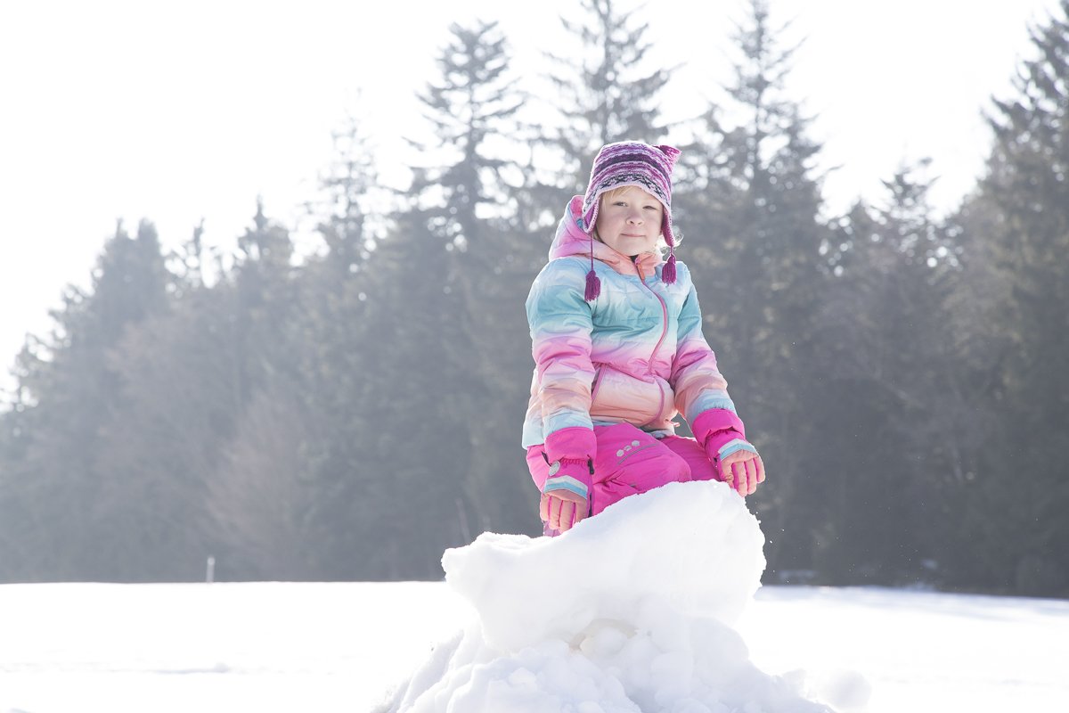Kind in bunter Winterkleidung sitzt auf einem Schneehaufen vor einem Wald