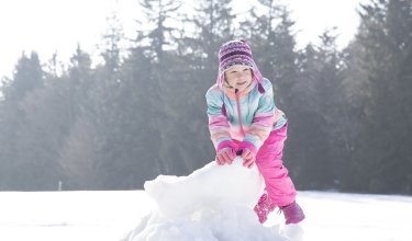 Kind in bunter Winterkleidung baut im Schnee mit Wald im Hintergrund