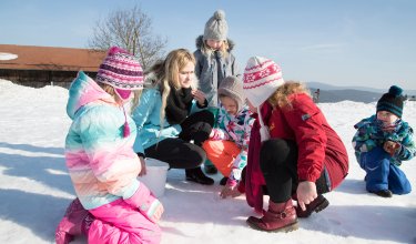 Gruppe von Kindern und einer Frau im Schnee, die etwas auf dem Boden betrachten, sonniger Wintertag