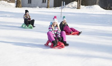 Drei Kinder rodeln auf einem schneebedeckten Feld vor einem Haus und Bäumen