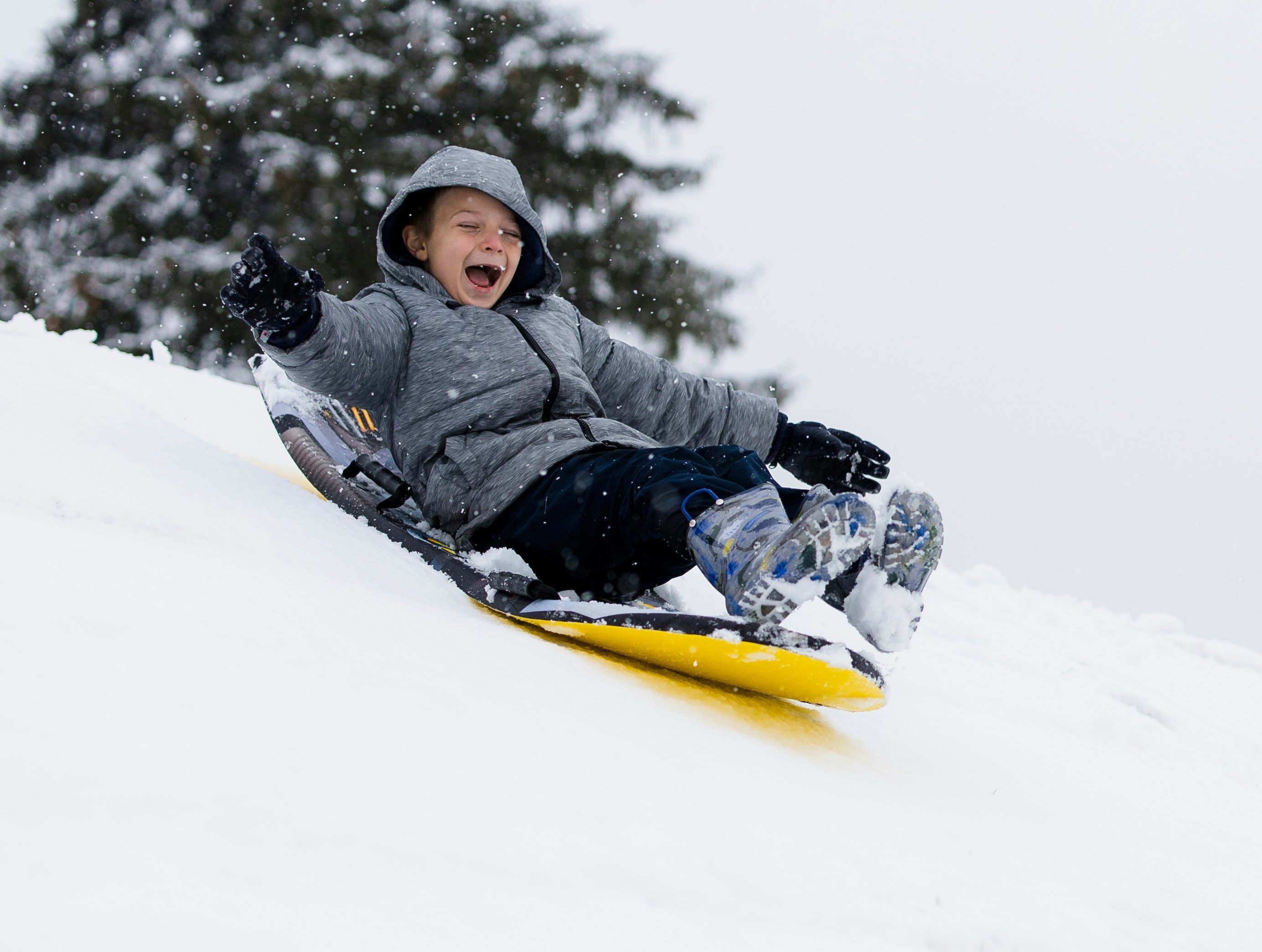 Kind in grauer Jacke rutscht lachend auf gelbem Schlitten einen schneebedeckten Hang hinunter