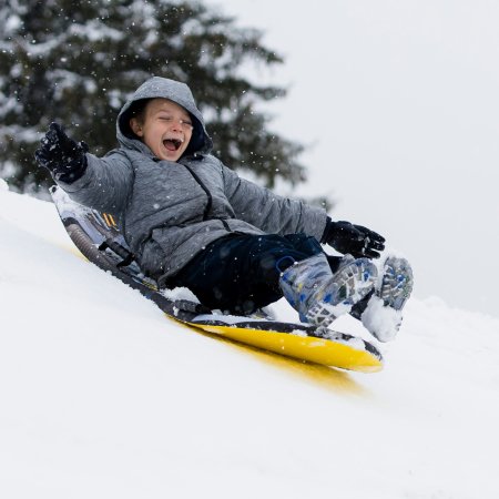 Kind in grauer Jacke rutscht lachend auf gelbem Schlitten einen schneebedeckten Hang hinunter