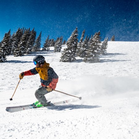 Skifahrer in bunter Jacke fährt den schneebedeckten Hang vor verschneiten Tannen unter blauem Himmel hinab