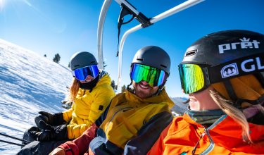 Drei Skifahrer in bunten Jacken und Helmen sitzen auf einem Skilift bei sonnigem Wetter