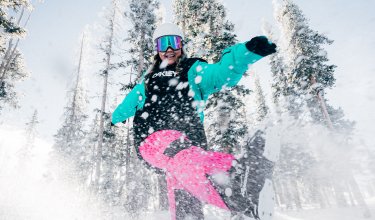 Person in bunter Winterkleidung kickt Schnee in einem verschneiten Wald unter blauem Himmel