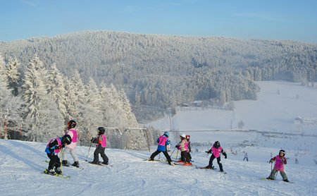 Kinder mit Skiausrüstung auf schneebedecktem Hang vor verschneitem Wald und blauem Himmel