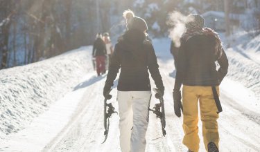 Zwei Personen gehen auf einem schneebedeckten Weg, eine trägt Schneeschuhe, die andere atmet sichtbar aus