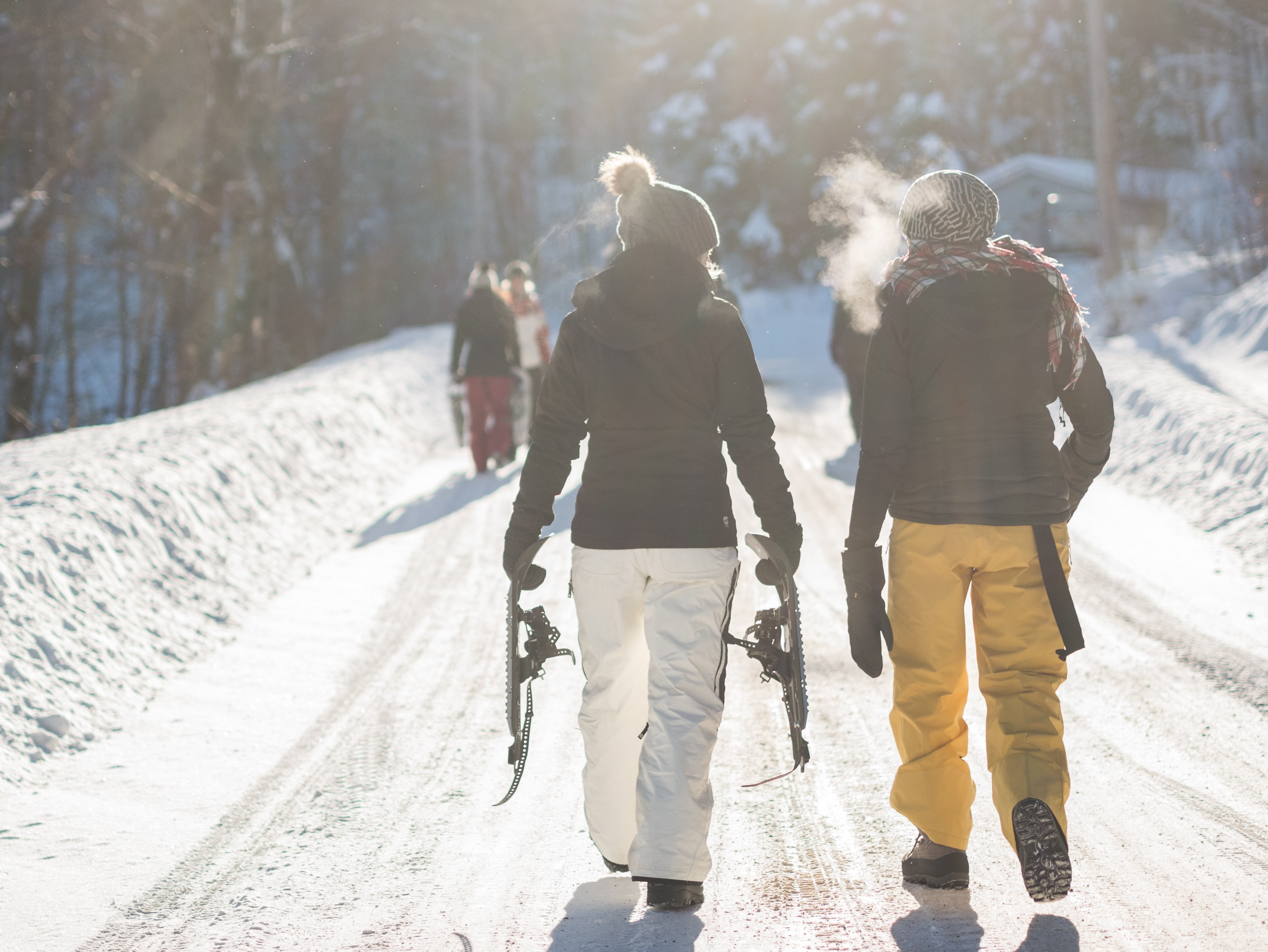 Zwei Personen gehen auf einem schneebedeckten Weg, eine trägt Schneeschuhe, die andere atmet sichtbar aus