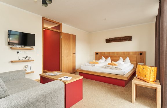 Helles Hotelzimmer mit Doppelbett, grauem Sofa, Fernseher an der Wand und orangener Tasche auf Holztisch
