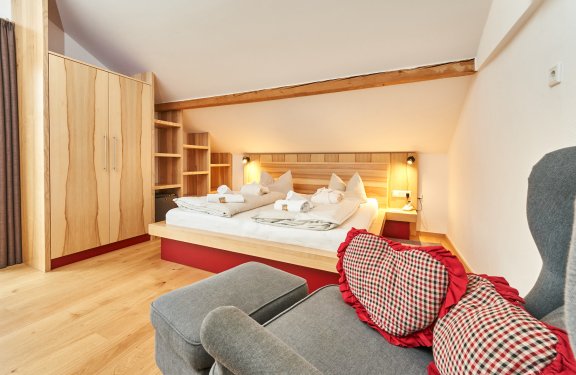 Gemütliches Schlafzimmer mit Holzmöbeln, Doppelbett und grauem Sessel mit karierten Herz-Kissen