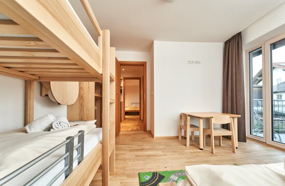 Helles Zimmer mit Etagenbett links, Tisch mit Stühlen rechts und Blick durch Tür zu weiterem Raum