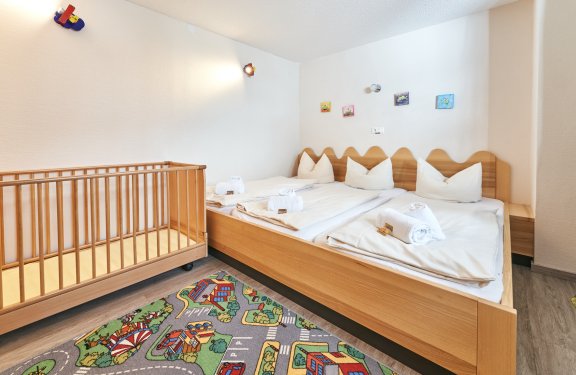 Kinderzimmer mit Holzgitterbett links, Doppelbett rechts und buntem Spielteppich auf dem Boden