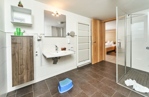 Helles Badezimmer mit Waschbecken, Spiegel, Holzschrank, offener Tür zum Schlafzimmer und Glasdusche
