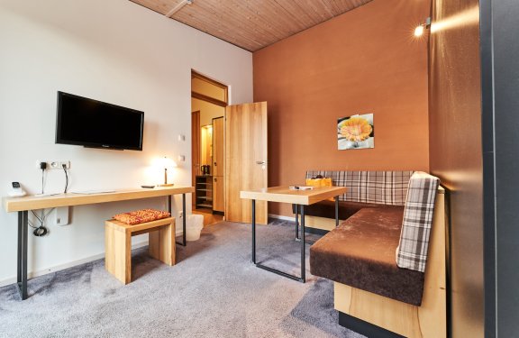 Gemütlicher Wohnbereich mit brauner Eckbank, Holztisch, Fernseher an weißer Wand und offenem Holztür