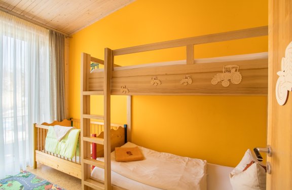 Kinderzimmer mit Etagenbett aus Holz vor gelber Wand und Babybett neben Fenster mit Vorhängen