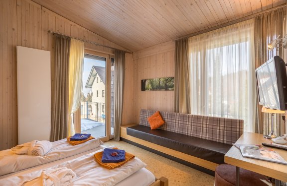 Helles Schlafzimmer mit Holzwänden, zwei Betten, Sofa, Fernseher und Balkon mit Blick auf Nachbarhaus