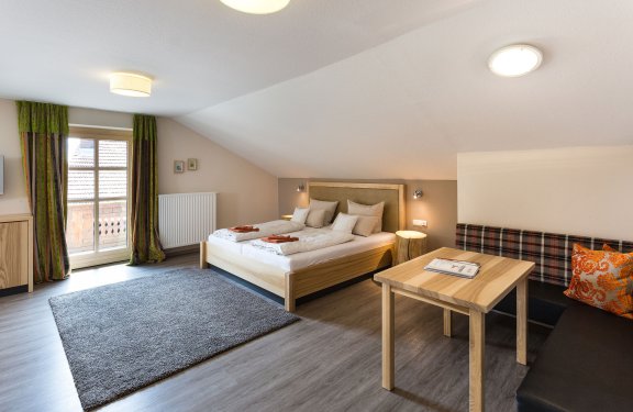 Helles Schlafzimmer mit Holzbett, Tisch, Sofa mit Kissen und Fenster mit grünen Vorhängen