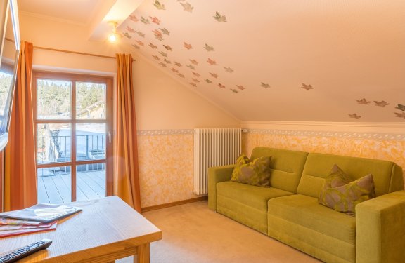Gemütliches Zimmer mit grünem Sofa, Holztisch und Fenster mit Blick auf Balkon und Wald