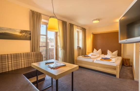Gemütliches Hotelzimmer mit zwei Betten rechts, Tisch und Sofa links, Fenster mit Vorhängen