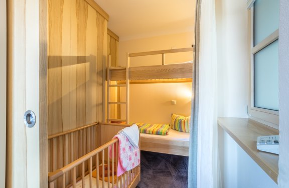 Kleines Zimmer mit Etagenbett und Babybett aus Holz, bunte Kissen und Decken, Fenster rechts