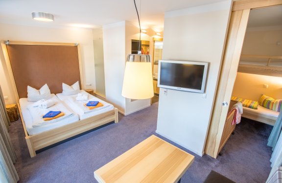 Helles Hotelzimmer mit Doppelbett, Fernseher an der Wand und angrenzendem Kinderzimmer mit Etagenbett
