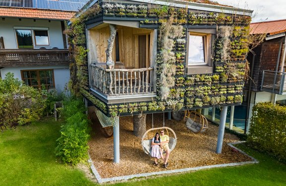 Haus mit begrüntem Holzfassade und Balkon, darunter sitzen zwei Personen auf einer Hängeschaukel