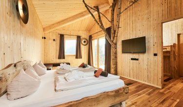Holzschlafzimmer mit Baumstamm im Raum, Doppelbett, TV an der Wand und Fenster mit Vorhängen