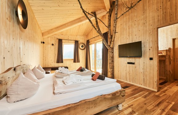 Holzschlafzimmer mit Baumstamm im Raum, Doppelbett, TV an der Wand und Fenster mit Vorhängen